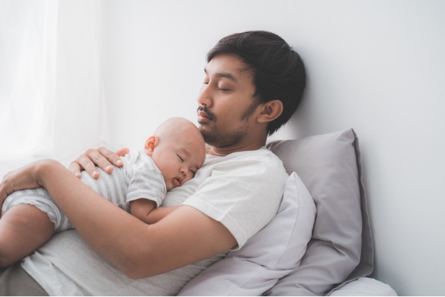 Un bébé dans les bras de son père, endormi tous les deux.